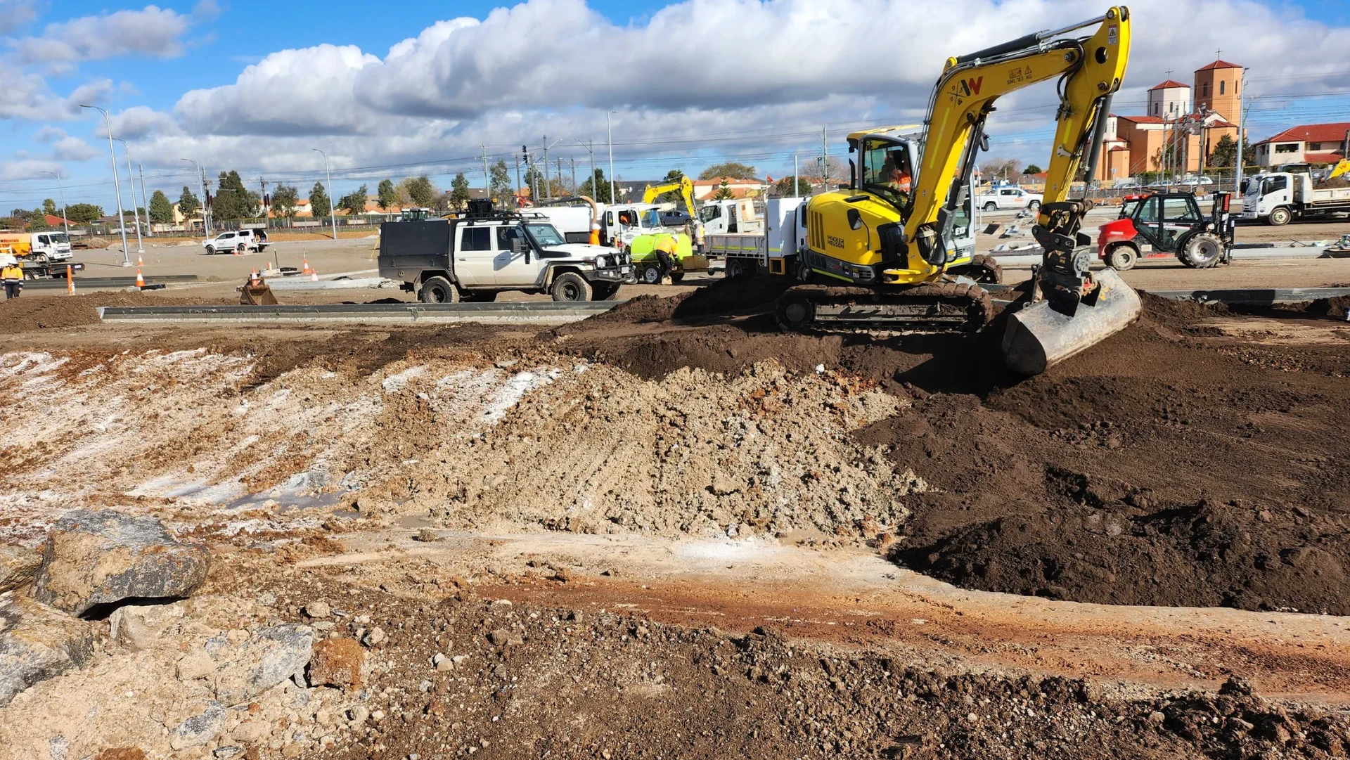 Excavator trimming subgrade beside newly placed kerb during formation and earthworks.